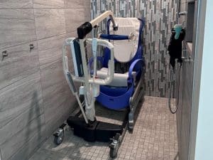 A blue and white shower wheelchair with padded seat and armrests is placed in a tiled accessible shower area with a handheld showerhead mounted on the wall.