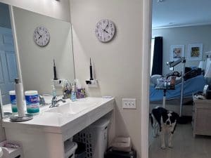 A bathroom with a white sink, mirror, two clocks showing different times, cleaning supplies, and toiletries. In the adjacent bedroom, a black and white dog stands near an unmade bed and a standing lamp.