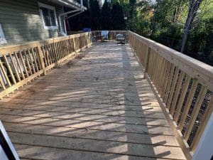 A long, wide wooden wheelchair ramp with railings on both sides leads from a house to the yard, surrounded by trees and sunlight. A dog is visible near the end of the ramp.