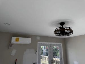 A ceiling fan with an industrial cage design is mounted on a white ceiling. Below it, double glass doors lead outside. An air conditioning unit is installed on a beige wall with several white paint patches.
