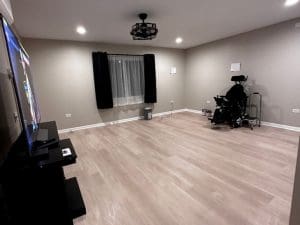A mostly empty room with wood flooring, beige walls, a large TV on a stand, a black curtain over a window, and a power wheelchair near the back corner beside a small table and trash can. Ceiling lights are on.
