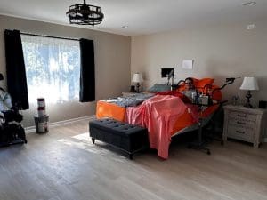 A spacious, sunlit bedroom with light wood floors, a large window with black curtains, and a bed covered in orange and pink bedding. A person is sitting up in bed using a tablet, with bedside tables and lamps on each side.