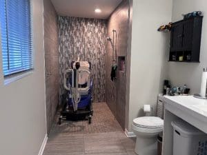 A modern bathroom features a roll-in shower with gray tile, a medical shower chair, a toilet, a countertop with a sink, storage cabinet, toilet paper holder, and a window with blinds.