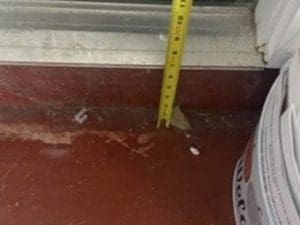 A yellow tape measure stands vertically against a red floor near a window sill, with a white bucket partially visible on the right side.