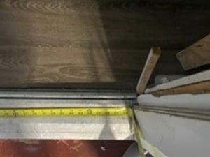 A yellow measuring tape stretched across a doorway with wood flooring and a white wall, showing a home improvement or construction setting. Pieces of wood and building materials are visible near the doorframe.