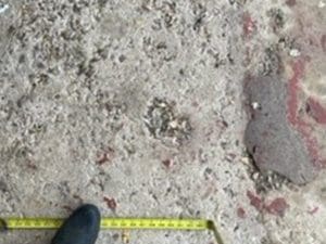 A yellow measuring tape is stretched on rough, uneven ground with scattered debris and a black shoe visible at the bottom left corner.