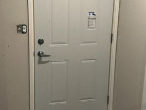 A closed, off-white door with four rectangular panels and two round metal handles is set in a beige wall. A light switch is visible on the left, and a label is attached to the top center of the door.