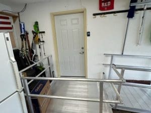 A metal wheelchair ramp leads to a closed white door in a garage. Gardening tools and equipment are hanging on the left wall, and a refrigerator stands in the left corner. Shelves and hooks are visible on the walls.