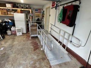 A metal wheelchair ramp is installed in a garage, leading up to a doorway. The garage contains a refrigerator, boxes, tools, and various household items along the walls.