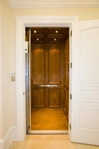 A residential elevator with wood-paneled walls and ceiling, recessed lighting, and an open white door. The elevator is empty and the interior looks clean and well-maintained.