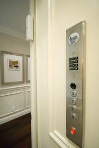 A close-up view of a home elevator control panel with a keypad, four numbered buttons, and emergency button. The elevator door is open, revealing a hallway with framed artwork on the wall.