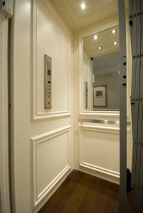 A small residential elevator with cream-colored paneled walls, wood flooring, a control panel with buttons on the left wall, a large mirror, and recessed ceiling lights.