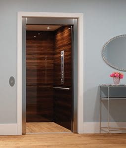 A modern home elevator with wood panel walls and open silver doors, set in a light-colored room with wood flooring, a glass table holding a pink flower arrangement, and a round mirror on the wall.