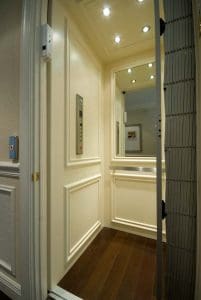 A modern residential elevator with cream-colored paneled walls, wood flooring, ceiling spotlights, and a metal accordion safety gate. A control panel and a mirror are mounted on one wall.