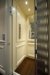 A well-lit residential elevator with cream-colored paneled walls, a mirrored back, a control panel, and a metal accordion safety gate partially open, situated on a dark wooden floor.