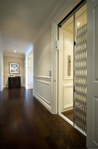 A residential hallway with dark wood floors and white trim features an open home elevator with a metal accordion gate. A small cabinet and framed artwork are visible at the end of the hallway.