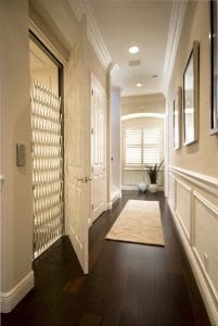 A hallway with dark wooden floors, white walls, framed pictures, and a beige rug leads to a window with plantation shutters. An open door reveals a home elevator with a metal accordion gate. A potted plant sits by the window.