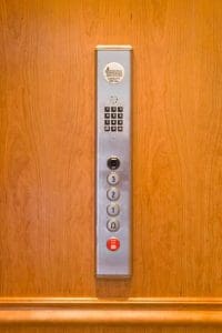 A vertical elevator control panel on a wooden wall with numbered buttons, a keypad, and an emergency stop button.