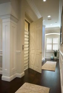 A hallway with dark wood floors, beige rugs, and a white door open to reveal a residential elevator with a metal gate. Sunlight streams through a window with white shutters at the end of the hall.