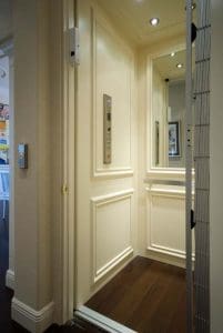 A small, elegant home elevator with cream-colored paneled walls, a control panel, a large mirror, and dark wood flooring. The elevator door is open, revealing part of a hallway outside.