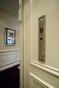 A close-up of an elevator control panel with buttons numbered 1 to 4, a red emergency button, and a keypad, mounted on a cream-colored wall inside a home with framed art visible in the background.