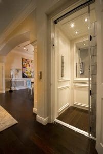 A modern home elevator with paneled white walls and a sliding metal gate is open in a hallway with dark hardwood floors, near an arched entry to a room with contemporary art and a dining table.