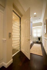A home hallway with dark wood floors and cream walls features an open elevator with a white safety gate. A beige rug leads to a window with shutters, next to a potted plant and a small table.