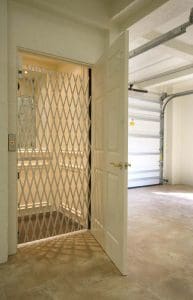 A residential elevator with a white accordion safety gate and an open white door is visible next to a garage space with tiled flooring and a partially open garage door.