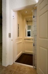 A home elevator with white paneled walls, wood flooring, a mirrored back wall, and a metal safety gate is open, showing the interior. The elevator is located next to a tiled hallway.