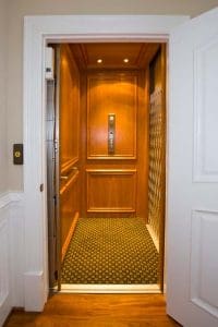 A home elevator with wooden paneled walls, a patterned carpeted floor, and an open white door. The control panel is visible on the back wall, and ceiling lights illuminate the interior.