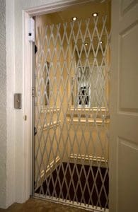 A residential elevator with an accordion-style metal gate is open, showing a well-lit interior with beige walls, paneling, and ceiling lights. An elevator button is visible on the wall outside.