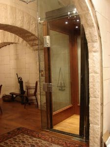 A small glass elevator with wooden interior and metal fixtures is built into a stone wall inside a room with wooden floors, arched stone doorways, and traditional furniture.
