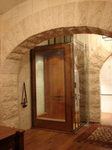 A modern glass-walled elevator with a wooden door is set into a stone archway in a rustic interior, featuring stone walls, wooden floors, and a decorative rug.