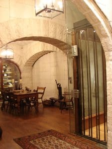 A room with stone arches, a wooden dining table and chairs, a glass door, and warm lighting. Wooden floors and a patterned rug add to the cozy, rustic atmosphere. A wine rack is visible in the background.
