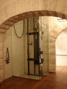 A modern glass elevator with black metal supports stands in a room with stone arches and beige tiled walls, featuring wooden flooring and soft, warm lighting.