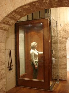 A woman stands inside a small, modern glass elevator with a wooden door, located in a room with arched stone walls and a wooden floor.