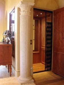A home elevator with wooden interior and black folding gate sits open next to a decorative stone column and wooden furniture in a warmly lit hallway.