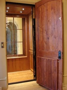 A wooden residential elevator with an open door reveals a paneled interior, wood flooring, and a large window, illuminated by ceiling lights and surrounded by beige walls.