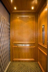 An empty, wood-paneled elevator with a patterned carpet floor, overhead lights, a metal accordion safety gate, and a control panel mounted on the right wall.