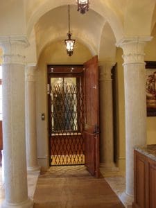 A home elevator with a wooden door and black accordion safety gate is located between two decorative stone columns under arched ceilings with warm lighting.