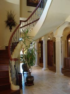 Elegant interior with a curved wooden staircase, decorative wrought iron railing, marble columns, beige tiled floor, and several potted plants. Natural light enhances the warm, inviting atmosphere.