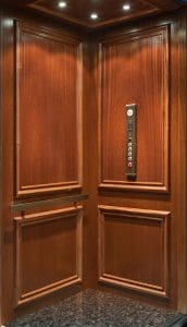 Wood-paneled elevator interior with a dark marble floor, built-in handrail, and a control panel featuring buttons and indicator lights mounted on the right wall. Four ceiling lights illuminate the space.