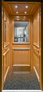 A wooden-paneled elevator with a dark, glossy floor, recessed ceiling lights, and a large mirror reflecting a bright hallway with windows and light-colored curtains.