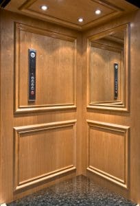 A modern elevator interior with wooden paneling, a mirrored wall, granite flooring, and a control panel with buttons and an emergency phone symbol. Ceiling lights illuminate the space.