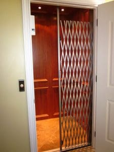 A residential elevator with wood-paneled walls and a metal accordion gate partially open, next to a closed outer door. The elevator call button is mounted on the wall beside the doorway.