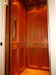 A modern residential elevator with rich wood-paneled walls, recessed ceiling lights, and a control panel with buttons on the left wall. The elevator door is partially open, revealing a tiled floor.