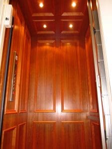 A wood-paneled elevator interior with recessed ceiling lights, decorative wall panels, and a control panel on the left side.