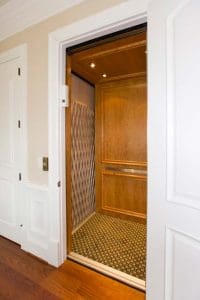 An open white door reveals a residential elevator with wood-paneled walls, a patterned floor, metal handrails, and a folding safety gate inside a well-lit, elegant home interior.