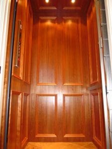 A wooden-paneled residential elevator interior with recessed lighting on the ceiling, control buttons on the left wall, and grooved panel designs on the walls and door.