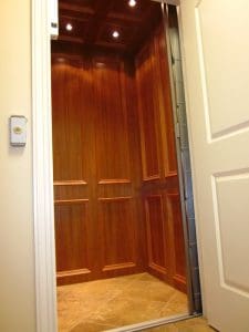 A home elevator with wooden paneled walls and a tiled floor is shown with the door open, revealing the illuminated interior. An elevator button is mounted on the wall outside.